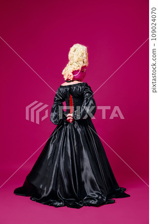 Cold revenge. Back view portrait of woman, in old-fashioned black dress holding colored knife behind her back against magenta background. Cold revenge. Back view portrait of woman, in old-fashioned black dress holding colored knife behind her back against magenta background. 109024070