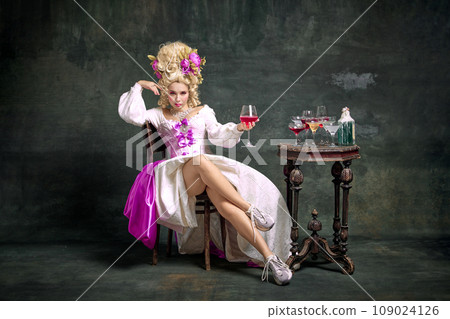 Serious princess, aristocratic medieval person in old-fashioned dress and modern shoes sitting holding cocktail against vintage background. Serious princess, aristocratic medieval person in old-fashioned dress and modern shoes sitting holding cocktail against vintage background. 109024126
