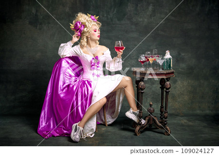 Charming queen, aristocratic medieval person in old-fashioned dress and modern shoes sitting holding cocktail against vintage background. Charming queen, aristocratic medieval person in old-fashioned dress and modern shoes sitting holding cocktail against vintage background. 109024127