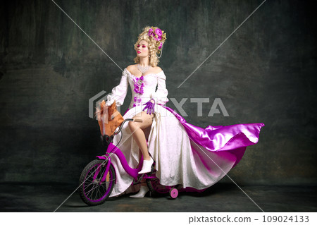 Serious woman in old-fashioned, aristocratic dress sitting, riding children's bike against vintage background. Concept of comparisons of eras. Serious woman in old-fashioned, aristocratic dress sitting, riding children's bike against vintage background. Concept of comparisons of eras. 109024133