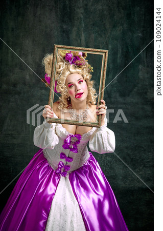 Woman dressed like classic historical character, in old-fashioned dress holding retro frame and grimacing looking at camera over vintage background. 109024144