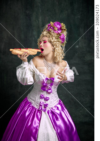 Portrait of young beautiful woman dressed like classical medieval person eating hot, delicious and huge American hot dog against vintage background. Portrait of young beautiful woman dressed like classical medieval person eating hot, delicious and huge American hot dog against vintage background. 109024173