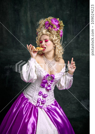 Historical portrait of young attractive woman in dress with corset eating delicious piece of pizza against vintage dark background. 109024223