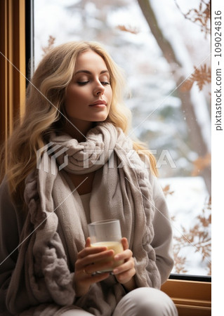 woman sits bundled up in scarf and jacket, sipping a warm drink while looking out at a snowy forest woman sits bundled up in scarf and jacket, sipping a warm drink while looking out at a snowy forest 109024816