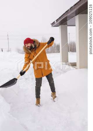 Young man clearing snow in his backyard village house with shovel. Remove snow from the sidewalk. 109025116