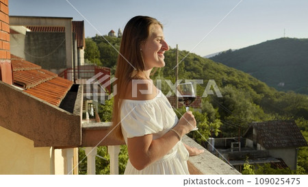 Young woman in dress enjoying glass of red wine on the balcony or villa terrace with view on sunset over mountains 109025475