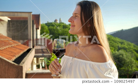 Smiling brunette woman with glass of red wine in hand, graces the balcony of a villa in the mountains at sunset. Spirit of travel, summertime, and holiday delight 109025497