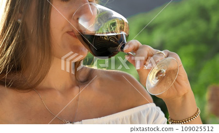 Closeup shot of elegant woman drinking red wine from glass against forest and mountain view 109025512