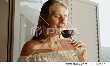 Portrait of beautiful brunette woman enjoying glass of red wine while standing in the doorway on villa terrace or balcony at sunset. Portrait of beautiful brunette woman enjoying glass of red wine while standing in the doorway on villa terrace or balcony at sunset. 109025546