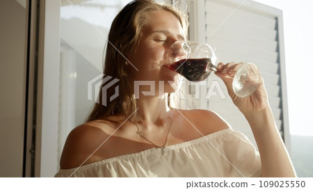 Portrait of brunette woman standing in a doorway on balcony and drinking red wine in sunset light 109025550