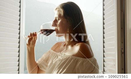 Beautiful young woman standing at open door on the terrace and drinks red wine from glass against sunset over the mountains 109025561