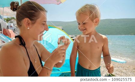 Young mother applying sunscreen spray on her baby son while relaxing on sea beach on a sunny day. Concept of healthcare, summer vacation, holidays with kids 109025575