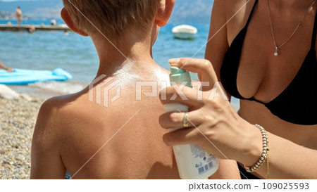 Closeup of caring mother sprays sunscreen spray on her son back and rubs it in his skin. Family holiday, summertime and vacation. Closeup of caring mother sprays sunscreen spray on her son back and rubs it in his skin. Family holiday, summertime and vacation. 109025593