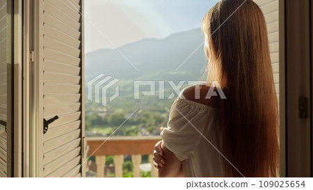 Young brunette woman opens window blinds at her villa and walking out on the terrace or balcony. Concept of summertime, enjoying mountain view, holiday and travel 109025654