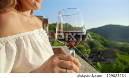 Young woman with glass of red wine gracefully held, merges with the magic of a mesmerizing sunset and mountain view from the villa balcony or terrace. Travel, summertime, and holiday serenity 109025684