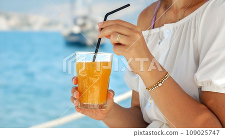 Closeup of woman sitting in the sea beach cafe and stirring orange juice with straw. Concept of summertime, relaxation, enjoying ocean. Closeup of woman sitting in the sea beach cafe and stirring orange juice with straw. Concept of summertime, relaxation, enjoying ocean. 109025747