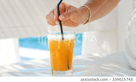 At a beach cafe, a woman stirs her orange juice with a straw, capturing the essence of summertime, relaxation, and the joy of seaside living At a beach cafe, a woman stirs her orange juice with a straw, capturing the essence of summertime, relaxation, and the joy of seaside living 109025748