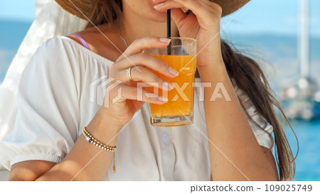 Closeup of elegant woman holding glass of orange juice and drinking it through straw while relaxing on wooden sea pier Closeup of elegant woman holding glass of orange juice and drinking it through straw while relaxing on wooden sea pier 109025749