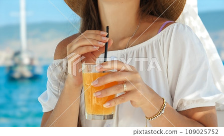 In a beach cafe gazebo on the pier, a brunette woman enjoys her orange juice with pure delight. Essence of summertime, holiday serenity, relaxation, and the call of travel 109025752