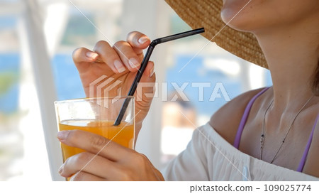 At a beachside cafe, a woman drinks orange juice, symbolizing summer holidays and coastal relaxation 109025774