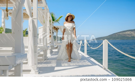 Smiling young woman, in a light dress and chic straw hat, walks on rustic wooden pier by the sea, infusing fashion with the allure of coastal bliss 109025866