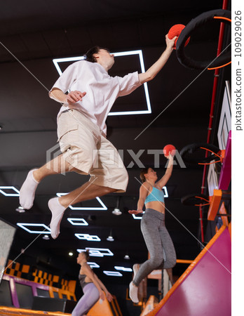 Energized young man enjoying amusement activities while jumping high on trampoline and hitting basketball hoop with ball in game club 109029026