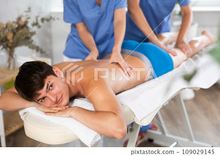 Relaxed young man having back massage therapy conducted by two masseuses Relaxed young man having back massage therapy conducted by two masseuses 109029145