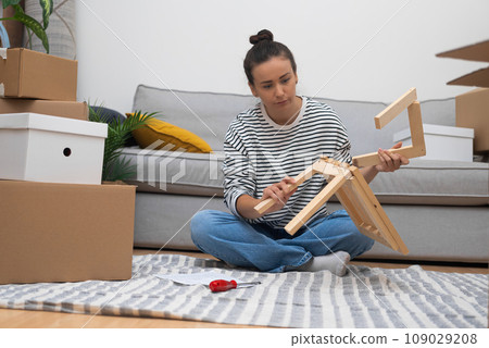 Young brunette woman sorts carton boxes of wooden furniture for new house sitting in spacious room with soft light with carpet waiting for lovely husband after work Young brunette woman sorts carton boxes of wooden furniture for new house sitting in spacious room with soft light with carpet waiting for lovely husband after work 109029208