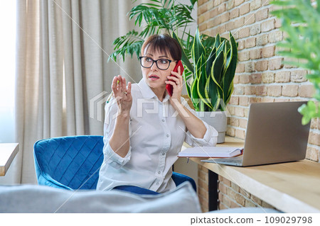 Middle-aged woman talking on mobile phone, sitting at desk at home with laptop 109029798
