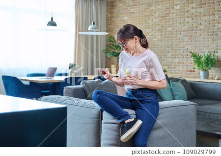 Middle aged woman with glass of water using smartphone sitting on sofa at home 109029799