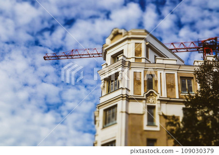 An old beige house, apartment building under construction on a city street. Construction crane machine over a roof and a blue cloudy sky. Restoration of an old building with stucco Roof reconstruction An old beige house, apartment building under construction on a city street. Construction crane machine over a roof and a blue cloudy sky. Restoration of an old building with stucco Roof reconstruction 109030379