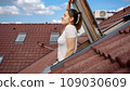A young woman gazes out of an open attic window with a red tiled roof in the background and breathes fresh air 109030609