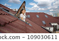 Happy smiling brunette woman opening attic window and breathing fresh air while looking on the cityscape with red tiled roofs 109030611