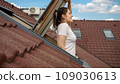 Woman smiling while looking out of an attic window under a red tiled roof and breathing fresh air 109030613