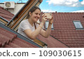 Young smiling woman looking out of the open attic window and making photos on her smartphone. Concept of tourism, holiday in Europe, travel and journey 109030615