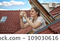 Young woman, with a bright smile, uses her smartphone to capture photos while looking out of an open attic window. Tourism, holidaying in Europe, and the spirit of travel and exploration 109030616