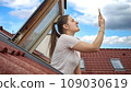 Young woman having issues with lost signal of a cellphone or smartphone looking out of the open attic window and trying to catch signal 109030619