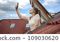 Young woman experiences signal issues with her cellphone as she peers out of an open attic window, attempting to regain a connection. Frustration of lost signal and connectivity problems 109030620