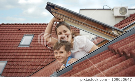 Happy smiling mother with son opens attic windows and looks outdoor. Concept of family happiness, parents with kids, moving to a new house. 109030647