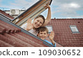 Smiling mother and her son open attic windows, gaze outside, and warmly wave towards the camera. Family happiness, parents with their children, and the excitement of moving to a new house 109030652