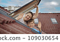 Happy mother and her son open attic windows, embrace the view outside, and warmly wave towards the camera. Family happiness, parents cherishing their children, and the exhilaration of settling into a 109030654