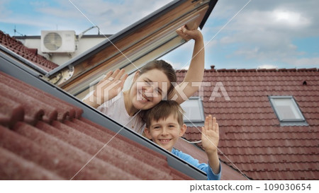 Happy mother and her son open attic windows, embrace the view outside, and warmly wave towards the camera. Family happiness, parents cherishing their children, and the exhilaration of settling into a 109030654