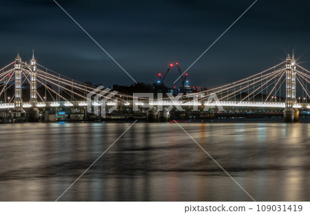 Nocturnal view of floodlit Albert Bridge from Chelsea Bridge with the reflection of the light in very calm water. 109031419