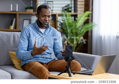 Serious concentrated man recording online video course, african american man using phone on tripod sitting on sofa in living room. Serious concentrated man recording online video course, african american man using phone on tripod sitting on sofa in living room. 109031806