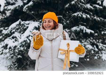 Smiling stylish 30s woman with present outside in the city park in winter. Young women with smartphone holding christmas present outdoors. Phone Communication. Hipster girl browsing Internet 109032921