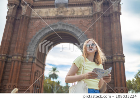 View of attractive young female tourist is exploring new city. Woman holding a paper map on central Woman making selfie against amazing architecture of the Arc de Triomphe gate in Barcelona View of attractive young female tourist is exploring new city. Woman holding a paper map on central Woman making selfie against amazing architecture of the Arc de Triomphe gate in Barcelona 109033224