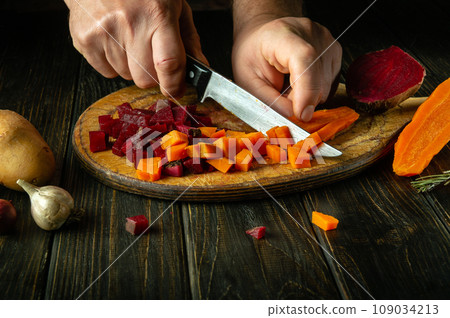 A cook uses a knife to cut boiled carrots to prepare a vegetable dish on the kitchen table. Venaigret cooking concept on dark background A cook uses a knife to cut boiled carrots to prepare a vegetable dish on the kitchen table. Venaigret cooking concept on dark background 109034213