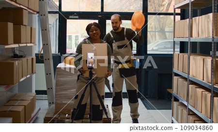 Warehouse marketing expert using smartphone placed on tripod to make ad campaign for social media platforms. African american influencer films herself in storage room showcasing her business Warehouse marketing expert using smartphone placed on tripod to make ad campaign for social media platforms. African american influencer films herself in storage room showcasing her business 109034860