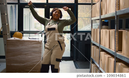 Chilled warehouse employee listening music enjoying herself while checking labels on cardboard boxes. Smiling worker wearing heaphones and dancing around while walking through storage hub rows Chilled warehouse employee listening music enjoying herself while checking labels on cardboard boxes. Smiling worker wearing heaphones and dancing around while walking through storage hub rows 109034861