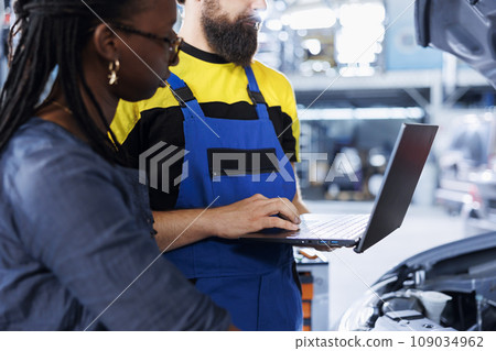 Mechanic in car service ordering new alternator for damaged vehicle using laptop. Technician next to customer looking online for components to replace old ones in malfunctioning automobile Mechanic in car service ordering new alternator for damaged vehicle using laptop. Technician next to customer looking online for components to replace old ones in malfunctioning automobile 109034962